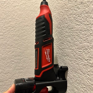 Puede incluir: Una herramienta el&eacute;ctrica Milwaukee roja y negra con una bater&iacute;a negra unida a un soporte de pared negro. La herramienta es una herramienta rotativa con una peque&ntilde;a punta unida al extremo.