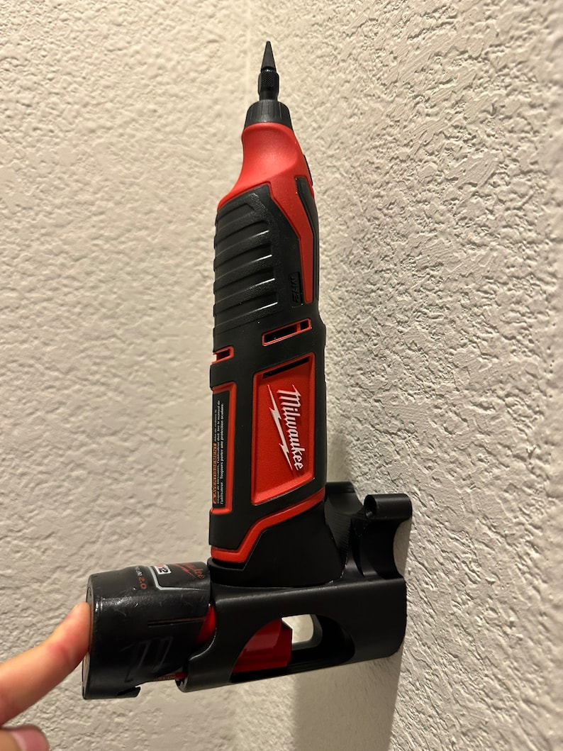 Puede incluir: Una herramienta el&eacute;ctrica Milwaukee roja y negra con una bater&iacute;a negra unida a un soporte de pared negro. La herramienta es una herramienta rotativa con una peque&ntilde;a punta unida al extremo.