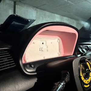 May include: A car dashboard instrument cluster housing featuring a black exterior frame and a light pink interior trim. The centre panel is white and rectangular, positioned behind a steering column with a yellow warning label attached.
