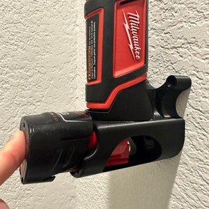 Puede incluir: Un soporte de herramienta el&eacute;ctrica negro y rojo con una herramienta el&eacute;ctrica Milwaukee unida. El soporte de la herramienta est&aacute; montado en una pared blanca. El soporte de la herramienta tiene una ranura para la bater&iacute;a y un gancho para la herramienta.