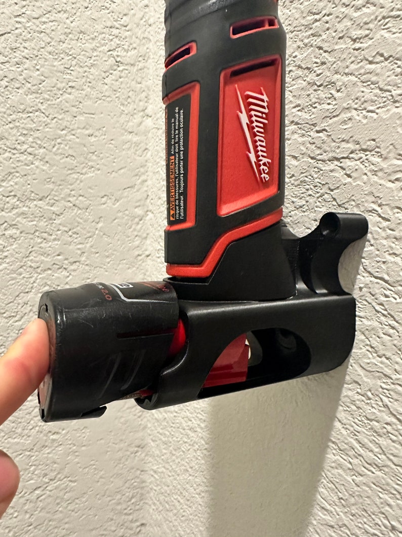 Puede incluir: Un soporte de herramienta el&eacute;ctrica negro y rojo con una herramienta el&eacute;ctrica Milwaukee unida. El soporte de la herramienta est&aacute; montado en una pared blanca. El soporte de la herramienta tiene una ranura para la bater&iacute;a y un gancho para la herramienta.