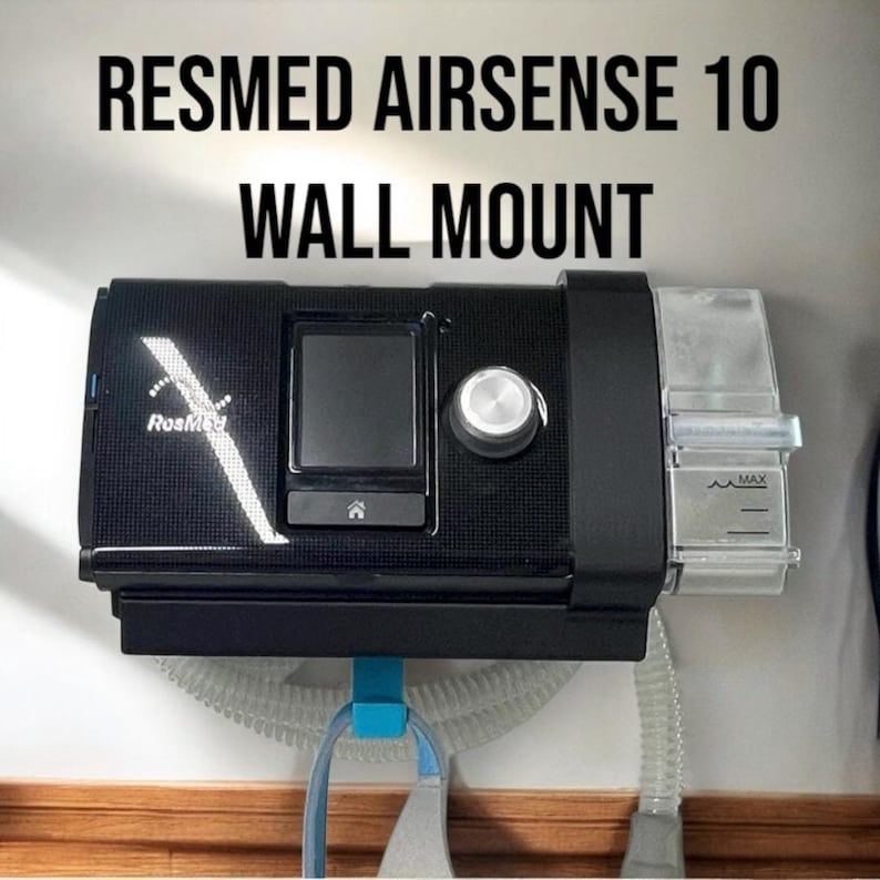 Puede incluir: Una m&aacute;quina CPAP ResMed Airsense 10 negra est&aacute; montada en una pared blanca. La m&aacute;quina tiene una pantalla digital y una perilla de control. Un tubo de pl&aacute;stico transparente est&aacute; conectado a la m&aacute;quina.