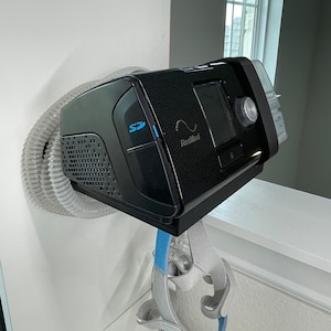 Puede incluir: Una m&aacute;quina CPAP negra con una manguera gris conectada a ella. La m&aacute;quina est&aacute; montada en una pared blanca. La m&aacute;quina tiene una peque&ntilde;a pantalla y un bot&oacute;n en la parte delantera. El texto "ResMed" es visible en la m&aacute;quina. Una m&aacute;scara CPAP gris est&aacute; colgando debajo de la m&aacute;quina.
