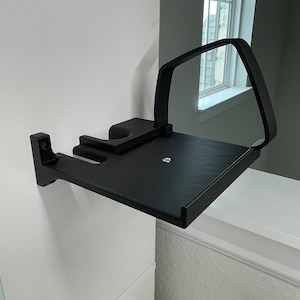 Puede incluir: Estante de pared negro con un brazo curvo para sujetar un dispositivo. El estante est&aacute; hecho de pl&aacute;stico y tiene un acabado mate.