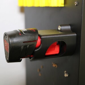 May include: A black and red battery holder for a power tool. The holder is mounted on a black surface. The battery is partially inserted into the holder.