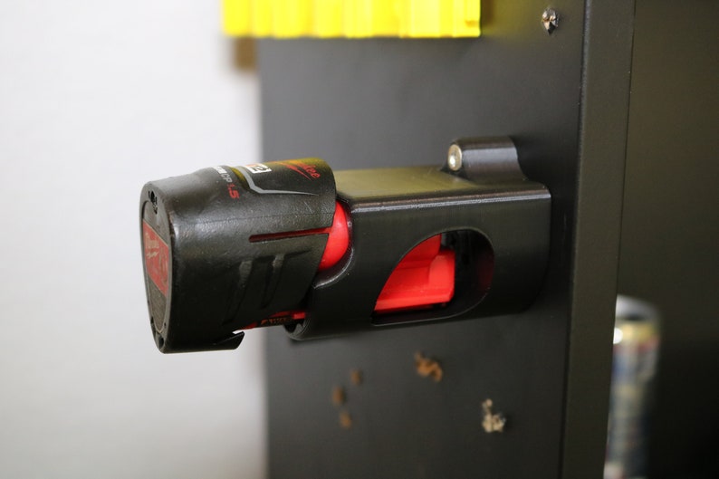 May include: A black and red battery holder for a power tool. The holder is mounted on a black surface. The battery is partially inserted into the holder.
