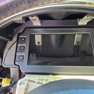 May include: A black Haltech digital dashboard display mounted in a vehicle instrument cluster. The unit features a rectangular screen surrounded by a black frame with three buttons on the left and three buttons on the right, including plus and minus symbols.