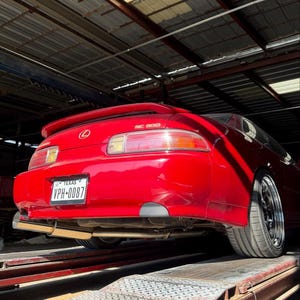 Pode incluir: Um cupê Lexus SC 300 vermelho visto de trás, com aerofólio, lanternas traseiras vermelhas e transparentes, ponteira de escapamento cromada e placa do Texas VPH-0087. O veículo está posicionado sobre uma plataforma elevatória de metal.