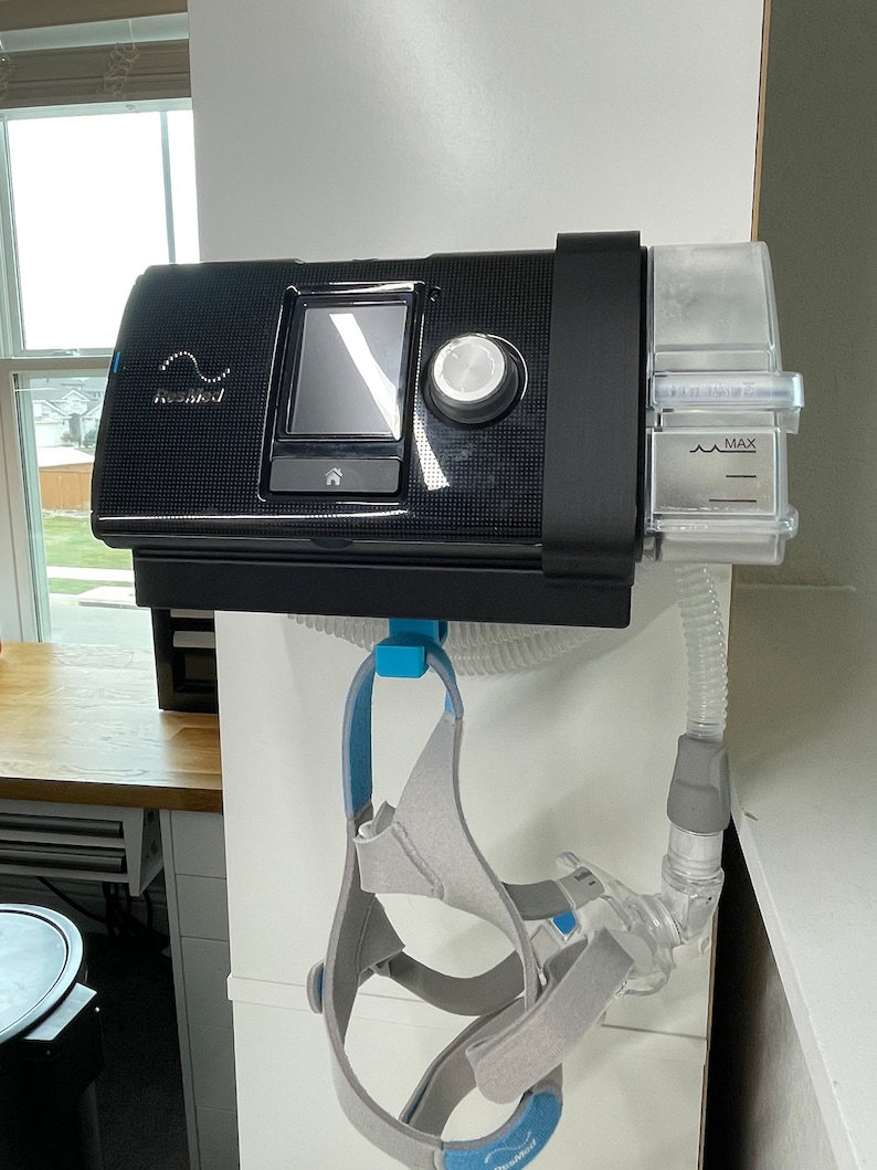 Puede incluir: Una m&aacute;quina CPAP negra con un tanque de agua transparente y una cinta para la cabeza gris colgando de ella. La m&aacute;quina est&aacute; montada en una pared.