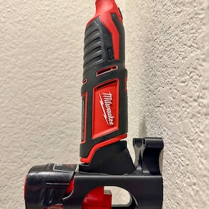 Puede incluir: Una herramienta el&eacute;ctrica Milwaukee negra y roja est&aacute; montada en un soporte de pared negro. La herramienta tiene un mango negro y rojo y un cuerpo negro y plateado. La herramienta est&aacute; etiquetada como "Milwaukee" y "M12".