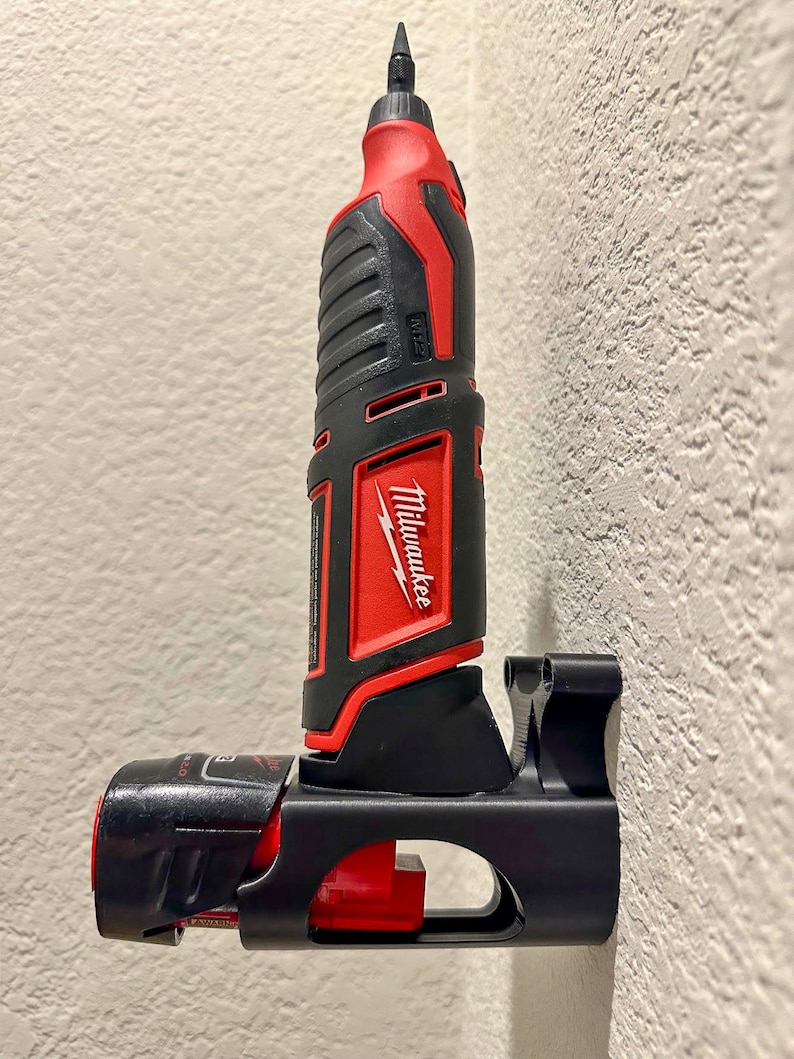 Puede incluir: Una herramienta el&eacute;ctrica Milwaukee negra y roja est&aacute; montada en un soporte de pared negro. La herramienta tiene un mango negro y rojo y un cuerpo negro y plateado. La herramienta est&aacute; etiquetada como "Milwaukee" y "M12".