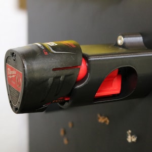 May include: A black and red power tool battery holder with a screw on the side. The holder is mounted on a black surface.
