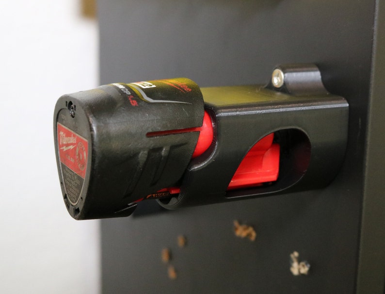 May include: A black and red power tool battery holder with a screw on the side. The holder is mounted on a black surface.
