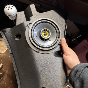 May include: A black speaker component with a round speaker and a yellow centre. The speaker is set in a textured black panel. The speaker is labelled "CS" and "Kicker".