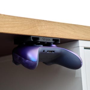 Op de afbeelding: Een paarse en blauwe Xbox-controller is gemonteerd onder een houten bureau met behulp van een zwarte plastic houder met het woord "Xbox" erop gedrukt.