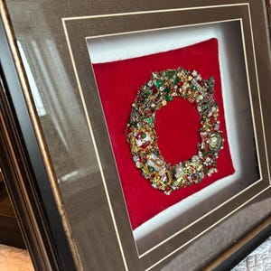 Pode incluir: Uma peça de arte de parede emoldurada apresentando uma guirlanda circular feita de joias vintage variadas, incluindo metal dourado, strass e miçangas coloridas, montada sobre um fundo de veludo vermelho brilhante dentro de uma moldura marrom escura com acabamento dourado.