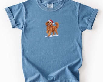 May include: A blue jean colored short-sleeve t-shirt featuring a graphic of a golden-brown curly-haired dog wearing a red and white Santa hat. The dog is standing on a small patch of white snow. The shirt has a crew neckline and rolled sleeves.