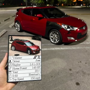 May include: A red Hyundai Veloster car with black accents is parked in a lot. A card held in front of the car displays the car's specifications, including the engine, horsepower, year, and top speed of 150 mph.