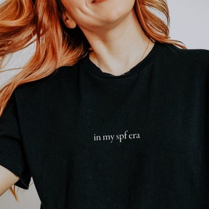 May include: A black oversized t-shirt with the text "in my sfera" printed in white on the front. The shirt is being worn by a person with red hair.