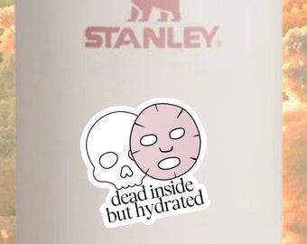 May include: White Stanley travel mug with a pink and white sticker that says "dead inside but hydrated". The sticker features a skull and a face mask.