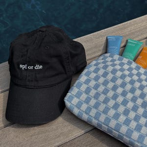 Puede incluir: Una gorra de béisbol negra con el texto "spf or die" bordado en la parte delantera. La gorra está sobre una superficie de madera junto a una bolsa de cuadros azules y blancos y tres tubos de protector solar.