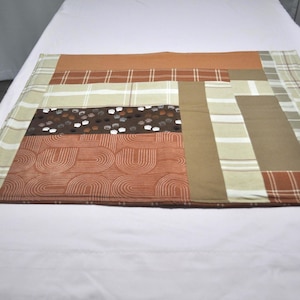 May include: A rectangular fabric placemat featuring a patchwork design of various earth-toned patterns. The textile includes sections of solid rust orange, tan, and brown, alongside plaid, striped, and a dark brown print with small white paw prints.