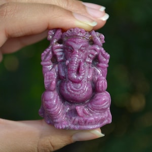 May include: A carved, magenta-colored statue of the Hindu deity Ganesha. The sculpture is held in a hand and features intricate details of the elephant-headed god in a seated position. The statue is approximately 2 inches tall.