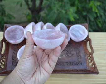 May include: A set of six small, pink rose quartz bowls on a wooden tray. The bowls are smooth and polished, and they have a natural, organic look.