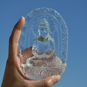 Könnte beinhalten: Eine klare, aufwendig geschnitzte Buddha-Statue aus Kristall. Der Buddha sitzt in meditativer Haltung, umgeben von einem bogenförmigen Rahmen. Die Statue wird vor einem strahlend blauen Himmel gehalten.