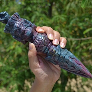 Puede incluir: Una daga tallada y ornamentada con un diseño detallado. La daga presenta un degradado de color de púrpura a turquesa, con intrincados patrones a lo largo del mango y la hoja. El objeto se sostiene contra un fondo verde borroso.