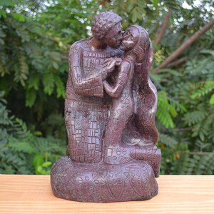 以下が含まれることがあります： キスをしているカップルの赤褐色の石の彫刻。彫刻は複雑なパターンとデザインで詳細に描かれています。