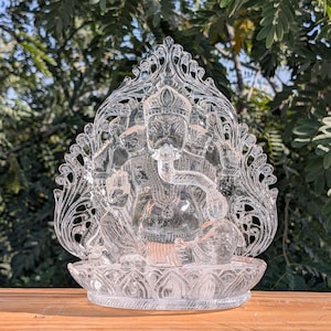 Könnte beinhalten: Eine klare, aufwendig geschnitzte Kristallskulptur der hinduistischen Gottheit Ganesha. Die Skulptur zeigt Ganesha auf einer Lotusblütenbasis sitzend, mit detaillierten Schnitzereien seiner Form und der umgebenden Ornamentik. Der Hintergrund zeigt üppiges grünes Laub.