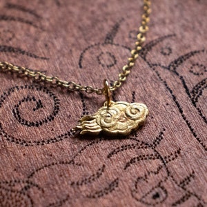 May include: A gold necklace with a cloud-shaped pendant. The pendant is detailed with swirls and is on a brown wooden surface.