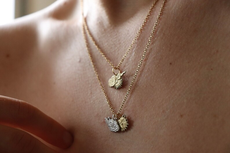 K&ouml;nnte beinhalten: Zwei goldene Halsketten mit zarten Ketten und Anh&auml;ngern. Die Anh&auml;nger sind beide Wolken- und Sonnenmotive, eines mit einem detaillierteren Design als das andere.