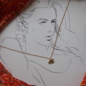 Op de afbeelding: Een gouden ketting met een klein hanger met een gestileerd zonontwerp. De ketting wordt gedragen door een vrouw in een lijntekening op een witte achtergrond.