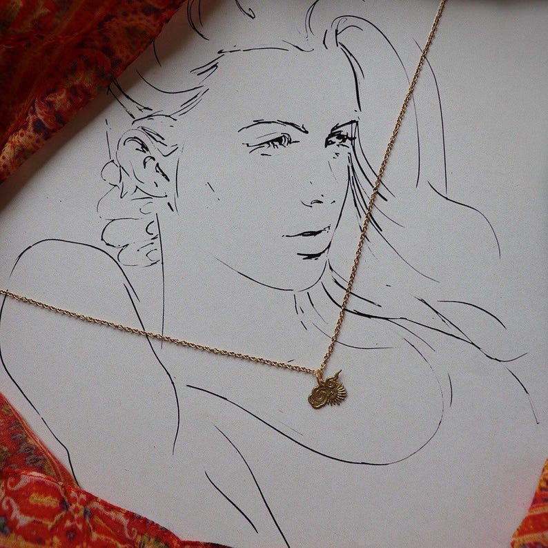 Op de afbeelding: Een gouden ketting met een klein hanger met een gestileerd zonontwerp. De ketting wordt gedragen door een vrouw in een lijntekening op een witte achtergrond.