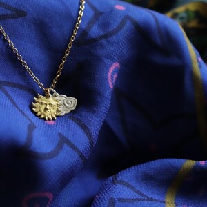 K&ouml;nnte beinhalten: Eine goldene Halskette mit einem Sonnen- und Wolkenanh&auml;nger. Die Kette liegt auf einem blauen Stoff mit einem rosa-wei&szlig;en Muster.