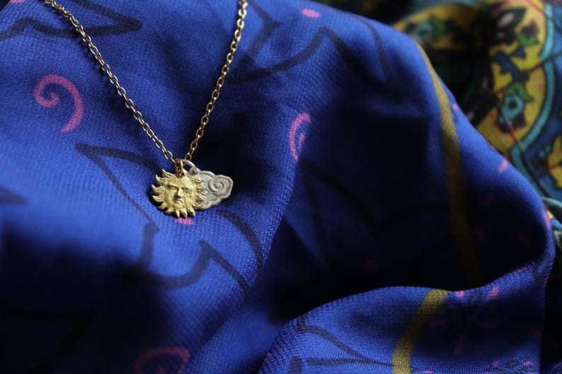 K&ouml;nnte beinhalten: Eine goldene Halskette mit einem Sonnen- und Wolkenanh&auml;nger. Die Kette liegt auf einem blauen Stoff mit einem rosa-wei&szlig;en Muster.
