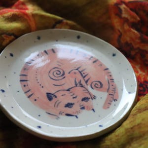 May include: A white ceramic plate with a pink and blue illustration of a curled up cat. The plate has a speckled glaze.