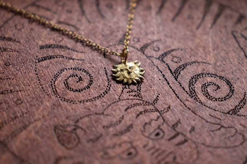 May include: A gold necklace with a sun pendant. The pendant is detailed with a face and rays. The necklace is on a brown surface with a pattern.