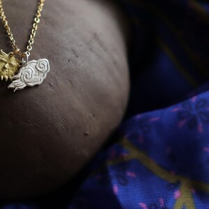 K&ouml;nnte beinhalten: Eine Halskette mit einem goldenen Sonnen- und einem silbernen Wolkenanh&auml;nger. Die Kette liegt auf einer braunen Oberfl&auml;che und einem blauen Stoff mit Muster.