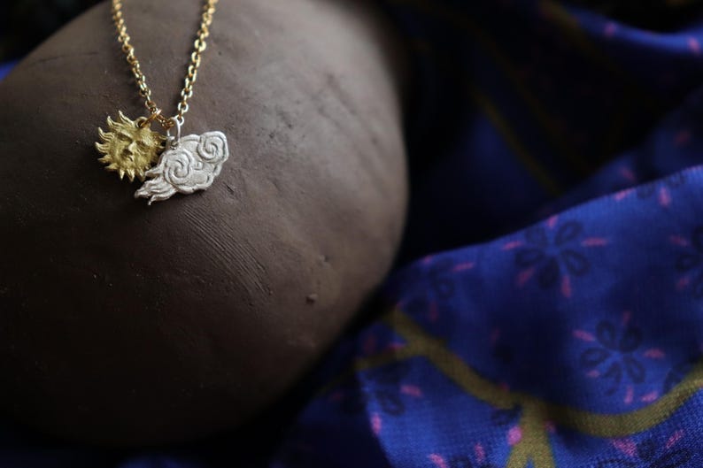 K&ouml;nnte beinhalten: Eine Halskette mit einem goldenen Sonnen- und einem silbernen Wolkenanh&auml;nger. Die Kette liegt auf einer braunen Oberfl&auml;che und einem blauen Stoff mit Muster.