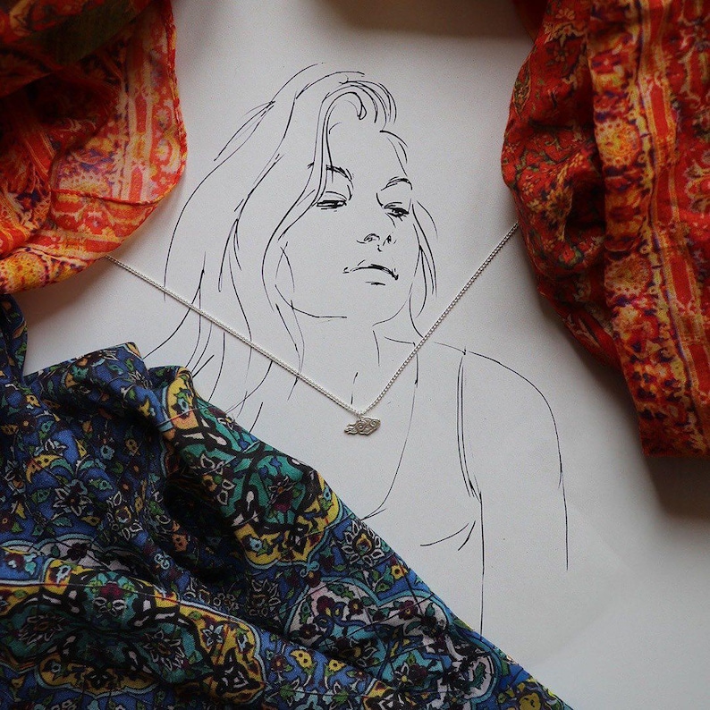 May include: A black and white line drawing of a person with long hair wearing a necklace with a small pendant. The drawing is on a white background with two patterned scarves on either side.