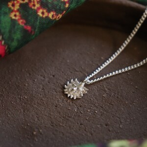 May include: A silver necklace with a sun pendant. The pendant is textured and has a detailed design. The necklace is lying on a brown surface.