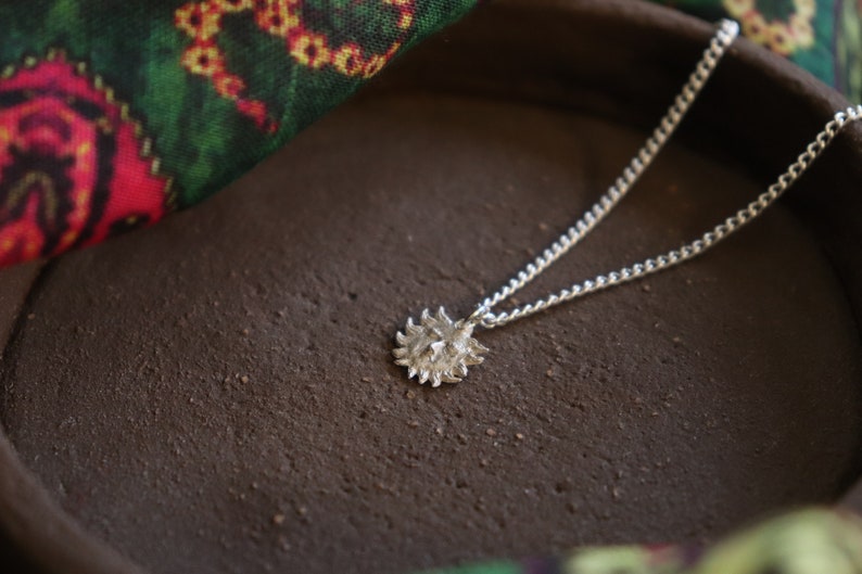 May include: A silver necklace with a sun pendant. The pendant is textured and has a detailed design. The necklace is lying on a brown surface.