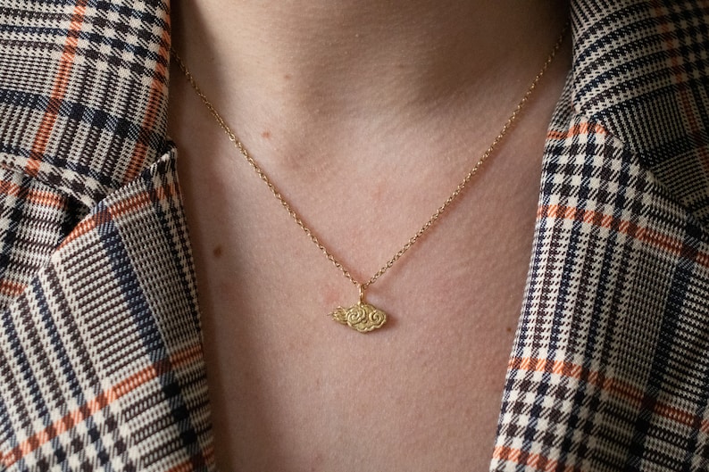 May include: A gold chain necklace with a small, detailed cloud pendant. The necklace is worn by a person wearing a plaid shirt.
