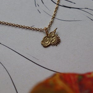 Op de afbeelding: Een gouden ketting met een bedel die een gestileerde zon en wolken voorstelt. De bedel is gemaakt van getextureerd metaal en heeft een delicaat ontwerp.