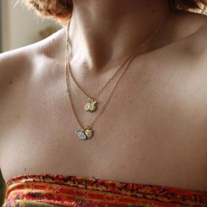 Op de afbeelding: Een persoon draagt een gelaagde gouden ketting met drie kleine bedels. De bedels zijn een zon, een halve maan en een klein blaadje.