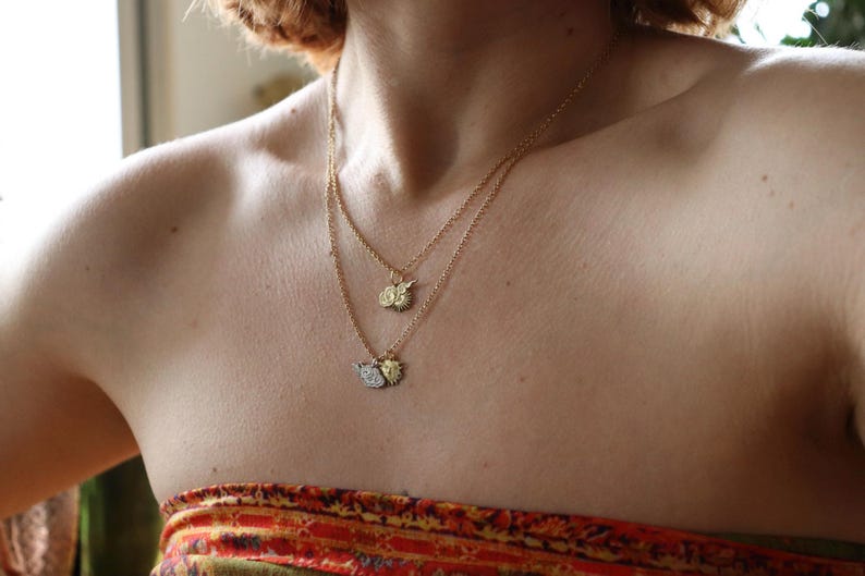 Op de afbeelding: Een persoon draagt een gelaagde gouden ketting met drie kleine bedels. De bedels zijn een zon, een halve maan en een klein blaadje.
