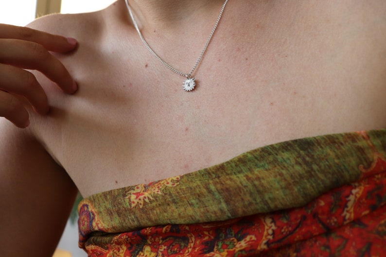 May include: A silver chain necklace with a small, silver sun pendant. The necklace is worn by a person with fair skin.